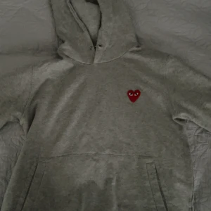 Grå hoodie Comme des Garçons Play - Grå hoodie från Comme des Garçons Play med det ikoniska röda hjärtat med ögon broderat på bröstet. Klassisk känguruficka, huva och långärmad modell. Mjuk bomullskvalitet som är perfekt för chill dagar. Helt ny och oanvänd, snöret till huvan följer med 