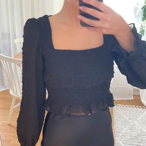 Svart blus med volang och puffärm - Supersnygg svart blus med fyrkantig ringning, puffig arm och smockad midja. Blusen har volangd nertill och är gjord i ett luftigt, srukturerat tyg. Som ny, använd 3ggr.🌟