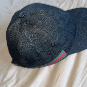 Svart keps från Gucci med GG-mönster - Snygg svart keps från Gucci med klassiskt GG-monogram över hela kepsen och grönt/rött band på sidorna. Justerbar rem baktill i skinn och detaljer i guld. Perfekt accessoar för att lyfta din outfit med lyxig känsla.