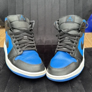 Nike Air Jordan 1 mid svart/blå - Säljer ett par Nike Air Jordan 1 High i svart och blått skinn med klassisk vit sula och svarta snören. Ikonisk Jordan-logga på sidan och tungan. Perfekta för dig som vill sticka ut med en tidlös sneaker med streetkänsla.