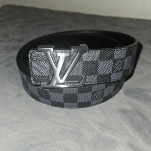 LV rutigt bälte i skinn - Snyggt bälte från LV med klassiskt rutigt mönster i svart och grått. Bältet har en stor silverfärgad LV-logga som spänne och är gjort i skinn. Perfekt accessoar för att lyfta din outfit med en exklusiv touch.