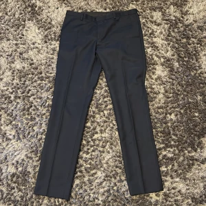 J.Lindeberg Kostymbyxor - Marinblå kostymbyxor från J.Lindeberg i slim fit-modell. Byxorna är tillverkade i 100% ull och har klassiska pressveck, bälteshällor och bakfickor med knappar. Perfekta för en clean och stilren look.