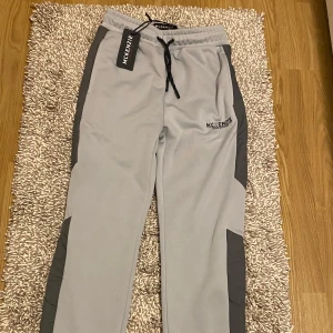McKenzie byxor - Snygga ljusgrå byxor från McKenzie med mörkgrå paneler längs sidorna. Byxorna har elastisk midja med svart snörning och logga på vänster lår. Perfekta för chill eller träning, tillverkade i mjukt syntetmaterial. Helt nya aldrig använda, storlek s