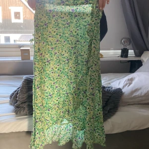 Grön blommig långkjol från Lindex - Säljer en flowy långkjol från Lindex i storlek 146/152. Kjolen är ljusgrön med blommigt mönster i lila, svart och gult. Den har resår i midjan och volang längst ner för extra vibe. Perfekt för dig som gillar färg och mönster!