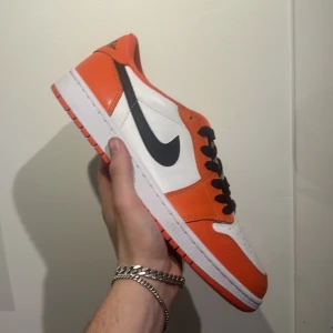 Nike Air Jordan 1 Low Orange/White - Nike Air Jordan 1 Low i orange och vitt med svarta detaljer och klassisk svart swoosh. Skon har svart snörning, perforerad tåbox och Air Jordan-logga på hälen. Tillverkad i skinn med platt vit sula och orange yttersula. Perfekt för dig som vill sticka ut med färg. 🇨🇳 🐼 