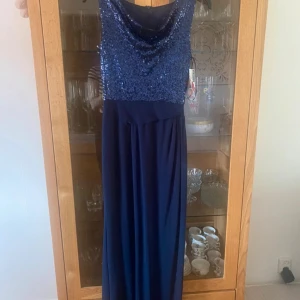 Marinblå långklänning med paljetter - Snygg marinblå långklänning med glittrig paljettöverdel och draperad halsringning. Kjolen är i ett mjukt, fallande tyg och klänningen är ärmlös. Perfekt för bal eller fest där du vill sticka ut med extra glamour.