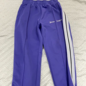 Lila Palm Angels mjukisbyxor - Snygga lila mjukisbyxor från Palm Angels med vita och svarta ränder längs ena benet. Byxorna har resår i midjan och logga tryckt på framsidan. Perfekta för en chill och sportig look.