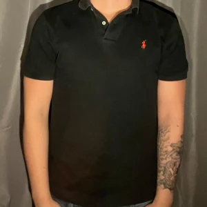 Svart pikétröja från Polo Ralph Lauren - Sommaplagg!🌞Klassisk svart pikétröja från Polo Ralph Lauren med röd broderad logga på bröstet. Tröjan har korta ärmar, krage och knappar vid halsen. Tillverkad i 100% bomull för en skön och stilren känsla.
