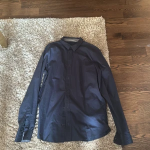 Mörkblå skjorta från H&M - Snygg mörkblå skjorta från H&M med klassisk krage och knäppning framtill. Skjortan har långa ärmar med manschett och är tillverkad i ett mjukt bomullstyg. Perfekt för dig som gillar en stilren och enkel look.