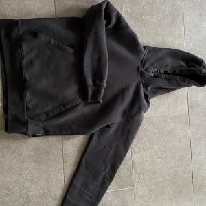 Svart Sail Racing hoodie med huva - Sparsamt använd Sail racing som jag köpte nypris 1200 kr på NK i Göteborg,    Strl S      Svart hoodie från Sail Racing med broderad logga på bröstet och stor känguruficka framtill. Tröjan har huva med snörning och är tillverkad i en mjuk bomulls- och polyestermix. Perfekt för dig som gillar en clean och stilren look.