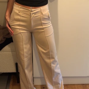 Beiga wide kostymbyxor med pressveck - Snygga beiga kostymbyxor halvt lågmidjade och raka, vida ben. Byxorna har pressveck framtill, hällor för bälte och klassiska fickor. Perfekta för en clean och stilren look. Materialet känns mjukt och har ett fint fall. Använda några gånger och lite smutsiga i benen men inget man direkt tänker på. 