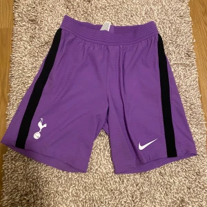 Lila Nike shorts Dri-FIT - Snygga lila shorts från Nike med svarta ränder längs sidorna och vita loggor. Tillverkade i Dri-FIT material som andas och passar perfekt för fotboll eller träning. Elastisk midja för skön passform och sportig look. Storlek s