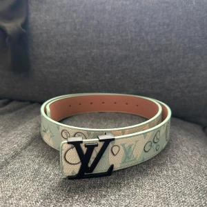 Louis Vuitton bälte med monogram - Snyggt bälte från Louis Vuitton med klassiskt monogrammönster i ljusa toner av beige och mintgrönt. Bältet har en svart LV-logga som spänne och är gjort i exklusivt skinn. Perfekt accessoar för att lyfta din outfit med en lyxig touch.