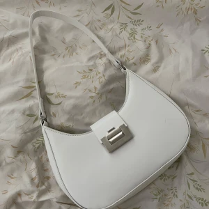 Vit axelväska med silverdetalj - Snygg vit axelväska i minimalistisk design med stilren silverfärgad låsdetalj framtill. Väskan har en enkel axelrem och ett huvudfack med dragkedja inuti. Perfekt för dig som gillar clean och modern look.