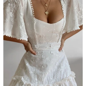Vit klänning från showpo - Superfin vit kortklänning med broderade blommor, volangkant och vida ärmar. Klänningen har en söt v-ringning med små bollar längs kanten och markerad midja. Perfekt för sommarens alla tillfällen och ger en romantisk vibe. Pytteliten fläck, märks dock inte!