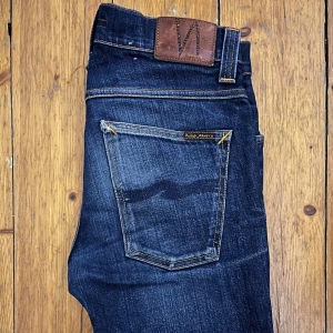 Mörkblå jeans från Nudie Jeans -  Mörkblå jeans från Nudie Jeans (modell Grim Tim) med riktig fett tvätt och orange sömmar. Jeansen harär tillverkade i högkvalitativt denim. Perfekta för en stilren look. Priset är inte hugget i sten.