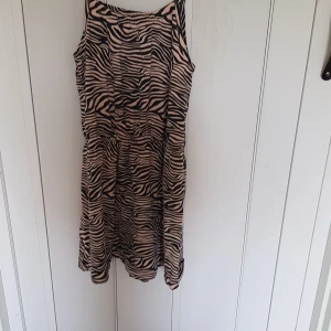 Zebramönstrad kort klänning Name It - Snygg kort klänning från Name It med zebramönster i svart och beige. Klänningen har smala axelband och resår i midjan för en skön passform. Perfekt för dig som gillar djurmönster och vill sticka ut lite extra.