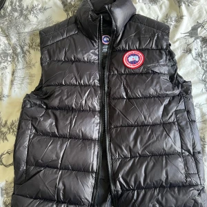 Svart dunväst från Canada Goose - Säljer en svart dunväst från Canada Goose med hög krage och dragkedja framtill. Västen har ett klassiskt Canada Goose-märke på bröstet och är quiltad för extra värme. Perfekt för kyliga dagar.