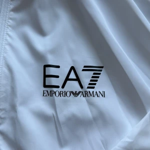 Vit dress från Emporio Armani EA7 - Snygg vit vindjacka från Emporio Armani EA7 med huva och halv dragkedja samt byxor. Jackan har svarta loggor på bröstet och ärmen, samt dragkedjeförsedda fickor. Lätt och sportig design, perfekt för dig som gillar stilrena och exklusiva plagg. Storlek S funkar även perfekt till M, använd ca 3 ggr och säljer pga storlek