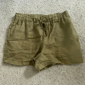 Olivgröna linneshorts med snörning - Snygga olivgröna shorts i linne med elastisk midja och snörning framtill. De har en avslappnad passform och två bakfickor för en chill vibe. Perfekta för varma dagar när du vill vara bekväm men ändå se fräsch ut.