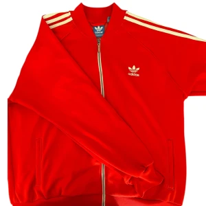 Röd Adidas bomberjacka med vita ränder - Snygg röd bomberjacka från Adidas med klassiska vita ränder på axlarna och broderad logga på bröstet. Jackan har dragkedja framtill, ribbade muddar och två fickor. Perfekt för dig som gillar sportig och retro stil.