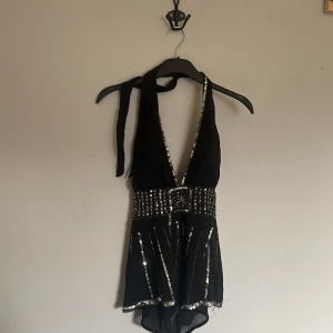 Svart halterneck topp med paljetter - Supersnygg svart halterneck topp med djup v-ringning och bredt paljettbälte i midjan. Toppen har glittriga silverdetaljer längs kanterna och nedtill, samt knyt i nacken. Perfekt för dig som vill sticka ut på festen!