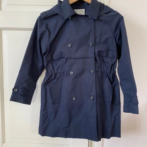 Mörkblå trenchcoat från Zara Girls - Snygg mörkblå trenchcoat från Zara Girls med huva, dubbelknäppning och knappar vid ärmslut. Jackan har dragsko i midjan för bättre passform och två fickor framtill. Fodret är randigt i blått och vitt, vilket ger en fräsch touch.