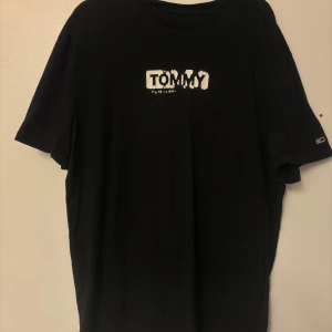 Svart t-shirt från Tommy Jeans - Svart t-shirt från Tommy Jeans med vit logga på bröstet och liten flaggdetalj på ärmen. Klassisk passform och rund halsringning, perfekt för en clean och stilren look. Tillverkad i mjuk bomull som känns skön mot huden.