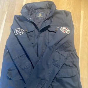 Mörkblå jacka från Henri Lloyd med patchar - Säljer en mörkblå jacka från Henri Lloyd med snygga patchar, bland annat en brittisk flagga och siffran 63 på ärmen. Jackan har flera fickor framtill, knappar och dragkedja. Perfekt för dig som gillar klassisk stil med sportig touch.