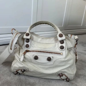 Vit Balenciaga handväska i skinn - Säljer en vit handväska från Balenciaga i mjukt skinn med coola metallknappar och dragkedjor i brons. Väskan har både handtag och axelrem, samt flera fack och detaljer som ger den en unik look. Perfekt för dig som gillar statement-accessoarer.