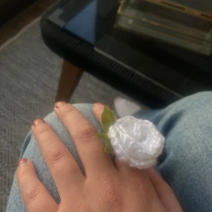Ring med virkad vit blomma - Unik ring med en handvirkad vit ros och gröna tygblad. Ringen har en justerbar metallbas i silverfärg och blomman är fluffig och mjuk, perfekt för dig som letar efter cool accessoar!!