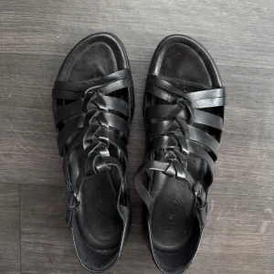 Svarta sandaler i skinn från ECCO - Snygga svarta sandaler från ECCO i mjukt skinn med flätade remmar och justerbart spänne vid ankeln. Platt sula och öppen tå ger en luftig känsla, perfekt för varma dagar. Klassisk och stilren design som passar till det mesta.