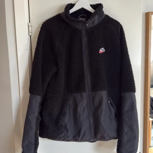 Svart teddyjacka från Nike - Svart teddyjacka från Nike med broderad logga på bröstet. Jackan har dragkedja framtill, två fickor och kontrastmaterial på ärmar och nederdel. Perfekt för kyliga dagar och riktigt skön att ha på sig.