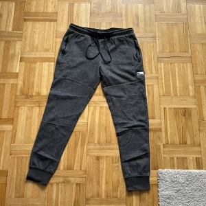 Mörkgrå mjukisbyxor Jack & Jones - Mörkgrå mjukisbyxor från Jack & Jones med snörning i midjan och dragkedjeförsedda fickor. Byxorna har ribbade muddar vid bensluten och en liten logotyp på benet. Perfekta för chill eller träning.