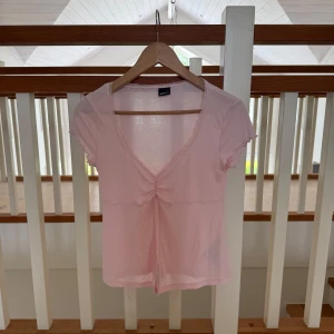 Ljusrosa v-ringad topp från Gina Tricot - Supersöt ljusrosa topp med v-ringning och rynkdetalj framtill från Gina Tricot. Korta ärmar med volangkant och spetsdetaljer längs halsringningen och framtill. Tunn och mjuk bomullskänsla, perfekt för vår och sommar.