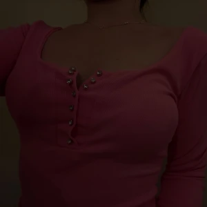 Rosa ribbad topp med knappar - Supersnygg rosa långärmad topp från Bik Bok med ribbad struktur och djup urringning. Framsidan har dekorativa silvriga knappar som ger en extra touch. Perfekt för dig som gillar en feminin och trendig stil.