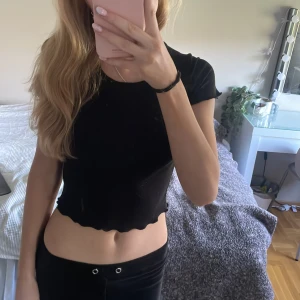 Svart croppad topp från Gina Tricot - Svart croppad topp från Gina Tricot med korta ärmar och vågig kant nertill och vid ärmslut. T-shirten är ribbad och har en tight passform som sitter snyggt på kroppen. Perfekt till jeans eller kjol för en enkel men trendig look.