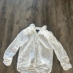 Vit linneskjorta från Polo Ralph Lauren - Snygg vit skjorta i linne från Polo Ralph Lauren med klassisk krage, långa ärmar och knappar framtill. Broderad marinblå logga på bröstet ger en clean och stilren look. Perfekt för dig som gillar avslappnad men ändå uppklädd stil.