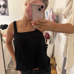 Svart ärmlös linnetopp med broderi - Svart ärmlös linnetopp med breda axelband och broderade detaljer upptill. Toppen har en luftig passform och fyrkantig ringning, perfekt för varma dagar. Snygg och enkel att matcha med jeans eller kjol.