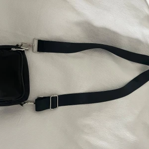 Svart axelväska med bred rem - En stilren svart axelväska med bred justerbar rem och två fack med silvriga dragkedjor. Väskan är kompakt och praktisk, perfekt för att bära det viktigaste. Tillverkad i syntetmaterial med metallspännen och detaljer.