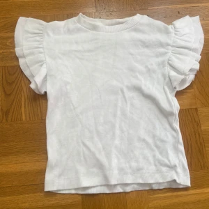 Vit ribbad topp med volangärm Gina Tricot - Superfin vit ribbad topp från Gina Tricot med korta volangärmar som ger en söt och trendig look. Perfekt till jeans eller kjol när du vill ha något enkelt men ändå extra. Mjuk och skön känsla mot huden.