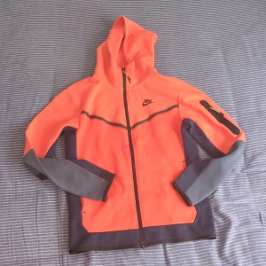 Nike orange hoodiejacka med dragkedja - Nike hoodiejacka i stark orange med mörkgrå detaljer och hel dragkedja. Jackan har huva, svarta Nike-loggan på bröstet och praktisk ficka med dragkedja på ärmen. Tillverkad i bomull och polyester, perfekt för en sportig och chill stil.