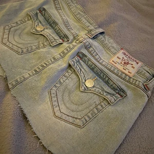 Ljusblå jeansshorts från True Religion - Säljer en ljusblå jeanskjol från True Religion med råa kanter och klassiska bakfickor med lock och silverfärgade knappar. Perfekta för sommaren! I storlek 28 (typ S)!