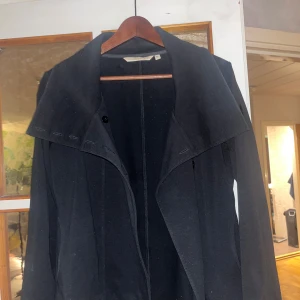 Svart trenchcoat från Tom Tailor - Säljer en svart trenchcoat från Tom Tailor med stor krage och bälte i midjan. Jackan har långa ärmar och en klassisk, stilren design som passar perfekt till höst och vår. Materialet känns mjukt och följsamt.