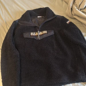 Svart teddyjacka från Napapijri - Mysig svart teddyjacka från Napapijri med halv dragkedja och broderad logga på bröstet. Jackan har hög krage och lång ärm, samt en praktisk ficka framtill. Perfekt för kyliga dagar.