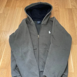 Mörk grön zip hoodie från Polo Ralph Lauren - Snygg grön zip hoodie från Polo Ralph Lauren med dragkedja och klassisk vit broderad logga på bröstet. Tröjan har huva, långa ärmar och är tillverkad i mjuk bomullsmix. Perfekt för en chill och stilren look.
