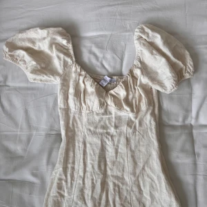 Linneklänning - Beige klänning i linnematerial. Den här puffärmar, markerade byst och sommar som ger en fin figur och har en slit. Går till vaderna och har en dragkedja på sidan samt resår på baksidan för att den ska sitta tight. Använd 1 gång. 