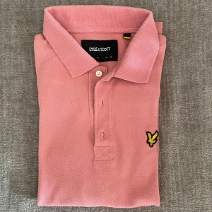 Pikétröja från Lyle & Scott - Snygg rosa pikétröja från Lyle & Scott med klassisk krage. Tröjan har det ikoniska gula fågel-loggan broderad på bröstet. Perfekt för dig som vill ha en snygg piké inför sommaren. Den är i perfekt skick och mycket sparsamt använd.