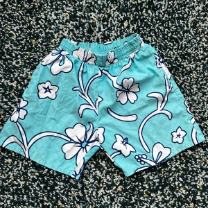 Turkosa blommiga badshorts - Snygga turkosa badshorts med stora vita blommor och marinblå konturer. Shortsen har elastisk midja och sidofickor, perfekta för strand eller pool. Lätta och snabbtorkande i polyester, med loose passform för chill vibe.