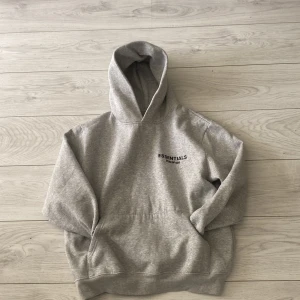 Grå hoodie från Essentials Fear of God - Säljer en stilren grå hoodie från Essentials Fear of God. Tröjan har en klassisk huva, ribbade muddar och tryck både fram och bak. Perfekt för en avslappnad och trendig look.